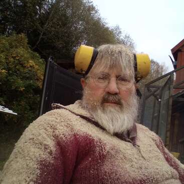 Ein bärtiger Mann mit Brille und gelben Ohrenschützern sitzt draußen, voller Sägemehl, wahrscheinlich nach der Arbeit mit dem Holz. Im Hintergrund sind Bäume und ein Gebäude zu sehen.