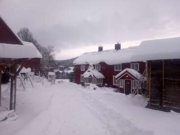 Eine verschneite Szene zeigt rote Holzhäuser und Scheunen, die unter einer dicken Schneedecke liegen. Der Himmel ist bewölkt, und die Landschaft wirkt friedlich, kalt und winterlich.