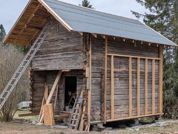 Esta imagem mostra uma cabana rústica de madeira em construção, com escadas encostadas nela. O telhado está parcialmente concluído e há materiais de construção espalhados ao redor.