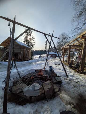 A rustic snowy outdoor setting with a fire pit, wooden structures, hanging grill, kettle, coffee mug, and chickens. The sun shines through the trees. Cozy atmosphere.