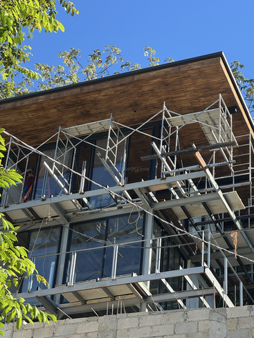 La imagen muestra un edificio moderno en construcción con andamios alrededor de las ventanas de cristal. La estructura cuenta con techos de madera, marcos metálicos, árboles y un cielo azul claro.
