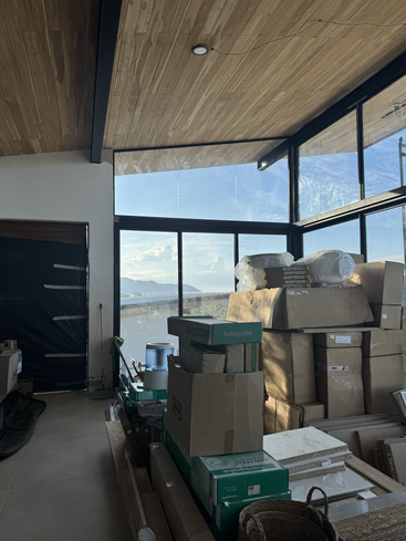 Una habitación con techo de madera y grandes ventanas, llena de cajas de cartón apiladas, materiales de construcción y herramientas, recibe luz natural y ofrece una vista panorámica del exterior.