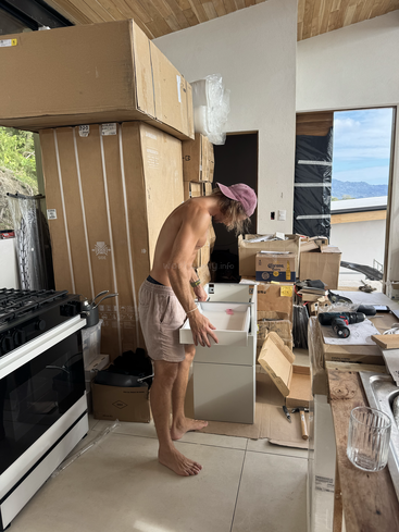 Un hombre sin camisa, vestido con pantalones cortos y una gorra rosa, monta muebles en una cocina abarrotada y soleada, rodeado de cajas, herramientas y artículos domésticos sin desembalar.