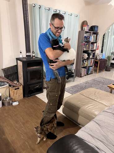 A smiling man in glasses is standing indoors, hugging two cats while a third cat weaves between his legs. The cozy room has bookshelves and a wood stove.