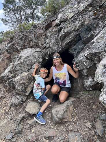 A woman and a child, both wearing headlamps, smile at the entrance of a rocky cave outdoors, ready for an adventure. Trees and rocks surround them.
