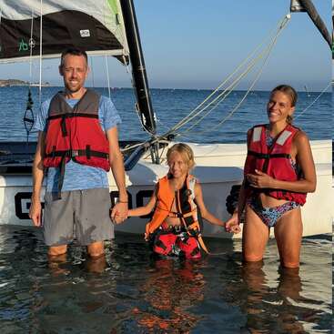 Eine fröhliche dreiköpfige Familie steht im flachen Wasser neben einem Segelboot, trägt Schwimmwesten und lächelt, während sie gemeinsam einen sonnigen Tag am Meer genießen.