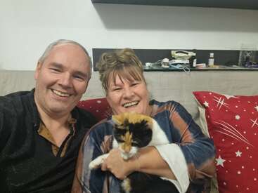 Um homem e uma mulher sorridentes sentam-se juntos em um sofá. A mulher segura carinhosamente um gato de chita. As almofadas vermelhas com estampa de estrelas dão um toque festivo à cena.