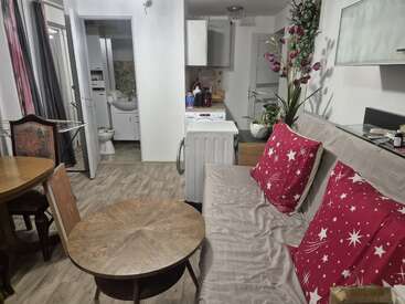 Este apartamento aconchegante possui uma pequena área de estar com sofá, almofadas vermelhas com estampa de estrelas, mesas redondas de madeira, cozinha compacta, máquina de lavar roupa e banheiro.