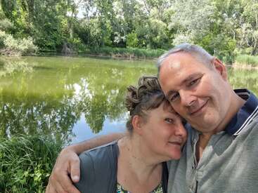 Una pareja se toma un selfie frente a un tranquilo estanque rodeado de frondosos árboles verdes, disfrutando de un día soleado y de su mutua compañía al aire libre.