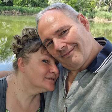 A couple takes a selfie in front of a peaceful pond surrounded by lush green trees, enjoying a sunny day and each other's company outdoors.