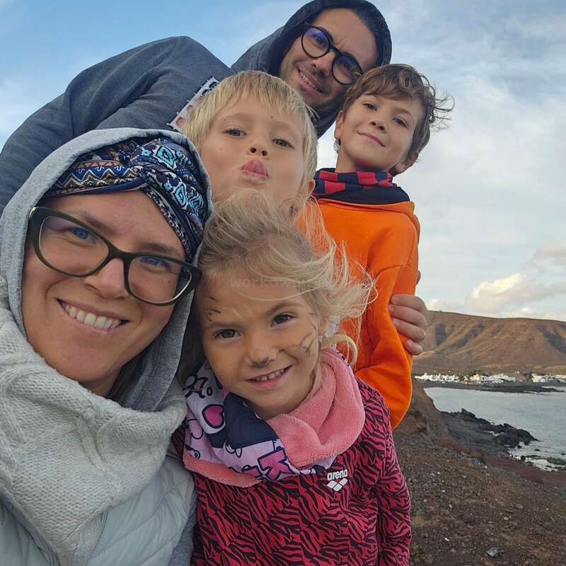 Une famille heureuse de cinq personnes pose en plein air au bord de la mer, souriant chaleureusement. Les enfants portent des vêtements colorés, l'un d'eux a le visage peint, avec les montagnes et le ciel en arrière-plan.