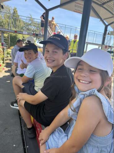 Three smiling children sit in a row at a miniature railway station, enjoying a sunny day out. Other people and a pedestrian bridge are visible behind them.