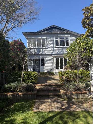 A charming two-story house with white siding, many windows, and a front garden featuring shrubs, trees, brick steps, and a sunny, clear blue sky.