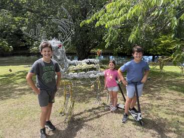 Three children smile outdoors beside a wire reindeer sculpture decorated with tinsel. Two ride scooters. Green trees and a pond provide a lush, sunny background.