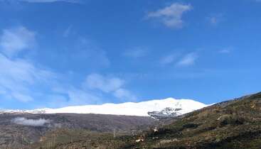 Uma paisagem de tirar o fôlego mostra montanhas cobertas de neve sob um céu azul intenso, com colinas verdes em primeiro plano, casas espalhadas e nuvens flutuando suavemente.