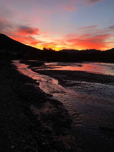 Um lindo pôr do sol pinta o céu com tons vibrantes de laranja e rosa, refletindo no rio tranquilo, cercado pelas silhuetas escuras das colinas e árvores.