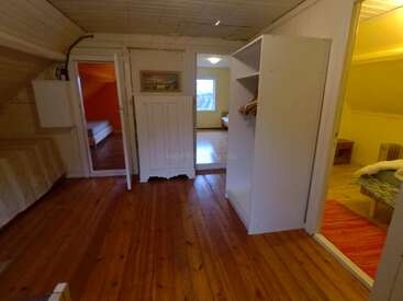 Cette image montre un couloir chaleureux avec un parquet, des murs lambrissés de blanc, trois chambres visibles, un mobilier sobre, des armoires ouvertes et un éclairage doux et chaleureux dans toute la pièce.