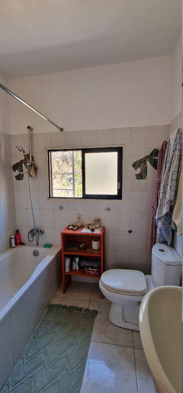 Este cuarto de baño cuenta con bañera, ducha, inodoro, lavabo, alfombra verde, ventana, estantería roja con artículos de aseo, toallas en ganchos y paredes de azulejos blancos.