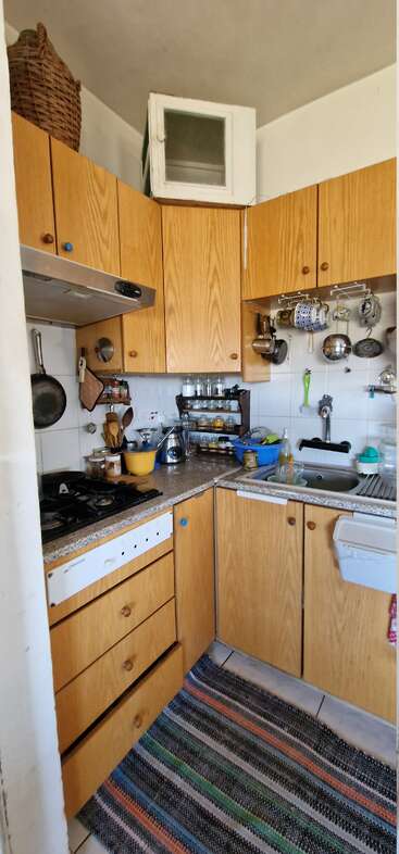 Una cocina compacta con armarios de madera, alfombra de colores, utensilios colgados, platos apilados, especias, tazas, un hornillo de gas y un fregadero junto a la encimera.