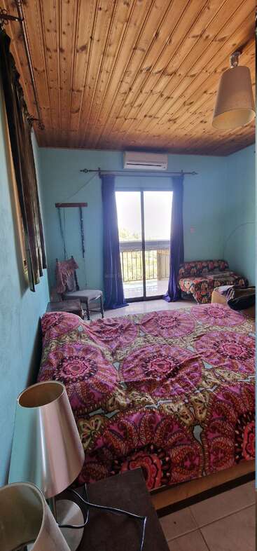 Un acogedor dormitorio con techo de madera, ropa de cama de colores, paredes azules, lámparas de cabecera, silla, sofá y puertas correderas de cristal que dan a un balcón con vistas.