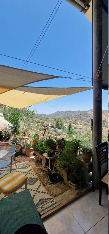 Un acogedor balcón iluminado por el sol cuenta con plantas en macetas, alfombras estampadas, velas de sombra, cómodos asientos y un pintoresco paisaje de montaña bajo un cielo azul brillante, creando un refugio tranquilo.