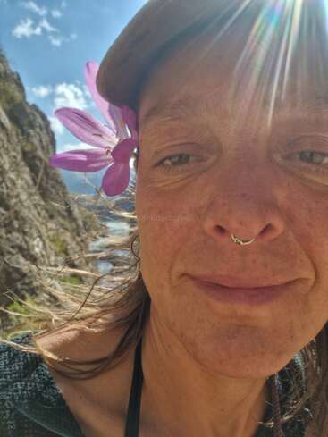 A close-up selfie of a smiling person outdoors, wearing a hat and septum ring, with a purple flower tucked behind their ear. Sunbeams light the scene.