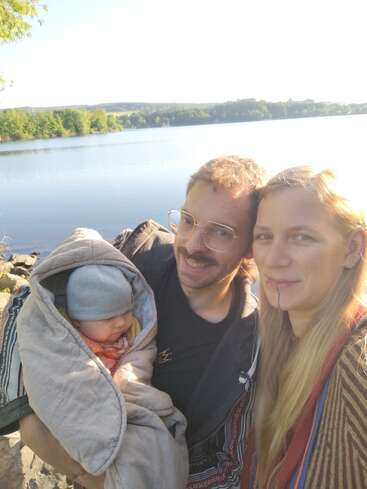 A happy family consisting of a man, woman, and baby pose outdoors by a tranquil lake. The baby is wrapped warmly. Trees and water shine brightly.