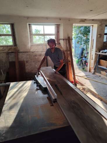 A man is working with a table saw in a sunlit, rustic workshop. Sunlight streams through the windows, illuminating sawdust and wooden boards scattered around.