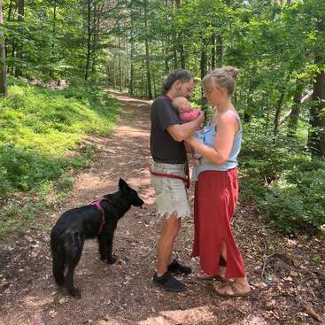 Ein Paar steht auf einem Waldweg und hält ein Baby im Arm. Sie schauen sich liebevoll an, während ein schwarzer Hund in der Nähe ist, umgeben von üppigem Grün.