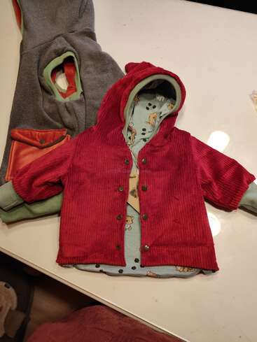 This image shows a small, red corduroy jacket with a hood, green cuffs, and animal print inner lining. Next to it is a grey garment.