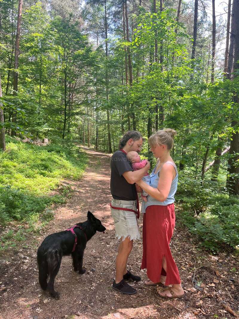 A couple stands on a forest path, holding a baby. They face each other lovingly, with a black dog nearby, surrounded by lush greenery.