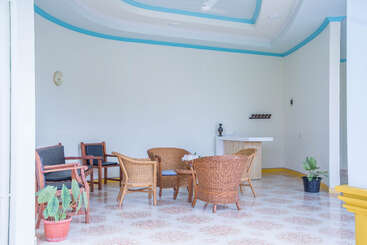 Das Bild zeigt einen hellen, luftigen Raum mit Korbstühlen und Holzmöbeln, zwei Topfpflanzen, einer Uhr und hellblau akzentuierten weißen Wänden und Decken.