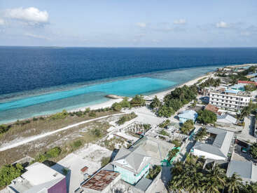 Dies ist eine Luftaufnahme einer Küstenstadt mit türkisfarbenem Wasser, weißem Sandstrand, verstreuten Gebäuden, üppigen grünen Bäumen und einem klaren blauen Himmel.