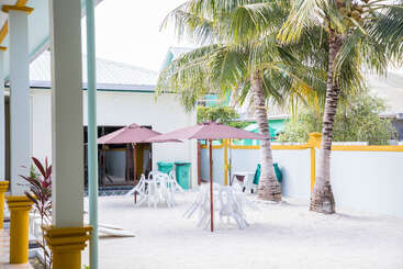 Ein sonniger Hof mit Palmen, weißem Sand, Plastiktischen und -stühlen, lilafarbenen Sonnenschirmen und farbenfrohen Gebäuden schafft eine entspannte tropische Atmosphäre, die sich perfekt für Freizeit und Versammlungen eignet.