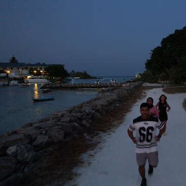 Trois personnes font leur jogging sur un chemin sablonneux longeant un rivage rocheux au crépuscule. Des bateaux flottent sur une eau calme, les lumières se reflètent et les arbres encadrent cette scène paisible.