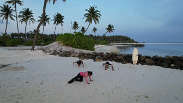 Three people are exercising on a tropical beach at sunrise, surrounded by palm trees, rocks, white sand, and a surfboard standing upright near the shoreline.