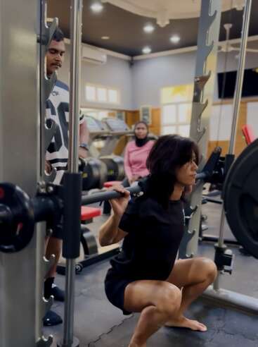 A woman is performing a squat exercise with a barbell in a gym. Two people observe her, one standing nearby and another sitting behind.