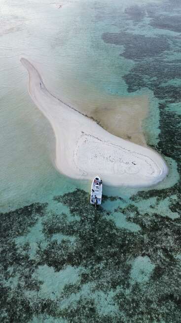 A small white sand island surrounded by shallow turquoise water, with a boat docked at its edge. One person walks along the narrow sandbar. Peaceful and isolated.