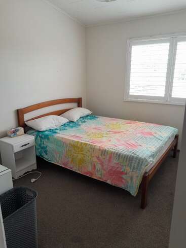 Un pequeño dormitorio con cama de matrimonio, colcha de flores de colores, dos almohadas, una mesilla de noche blanca, una caja de pañuelos, contraventanas y un cesto para la ropa sucia.