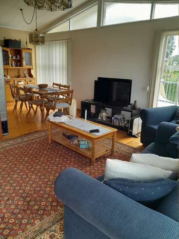 This cozy living room features blue couches, a patterned rug, a wooden coffee table, TV, dining area, chandelier, large windows, books, and a china cabinet.