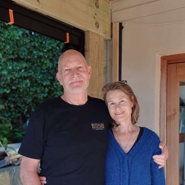Una pareja sonriente permanece unida en un ambiente acogedor de interior y exterior. El hombre lleva una camisa negra, mientras que la mujer lleva un jersey azul. Ambiente cálido.