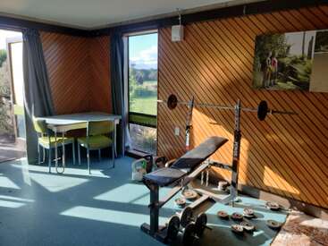 Une petite salle de sport aux murs lambrissés comprend un banc de musculation, des haltères, des barres et une table avec deux chaises vertes. La lumière du soleil illumine l'espace.