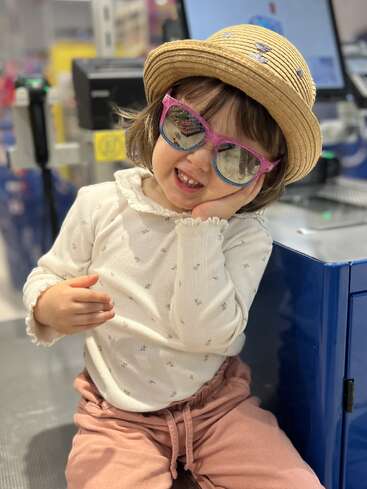 A young child wears a straw hat and pink sunglasses, smiling playfully. Dressed in a white shirt and pink pants, sitting indoors near a blue counter.