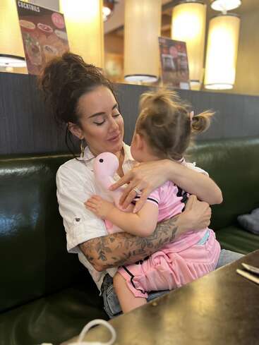 A woman lovingly hugs a young child on her lap in a restaurant booth. The child holds a plush toy. Warm, intimate moment, surrounded by soft lighting.