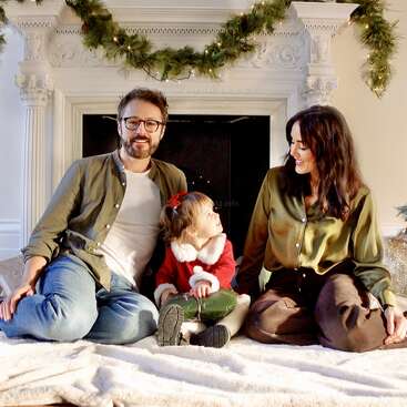 Uma família de três pessoas está sentada alegremente em frente a uma lareira decorada com guirlandas, compartilhando sorrisos e calor humano, vestida com roupas de inverno festivas e aconchegantes.