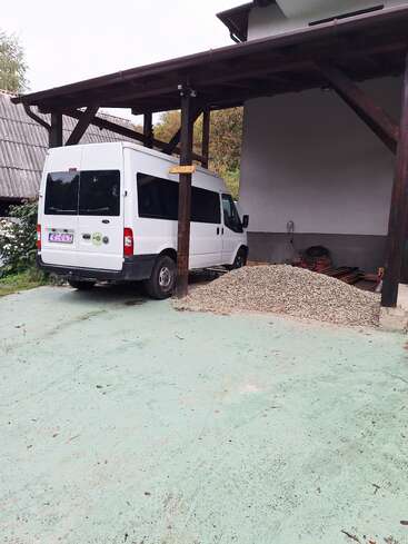 Une camionnette blanche est garée sous un abri de voiture en bois attenant à une maison. Un tas de gravier et divers matériaux de construction se trouvent à proximité.