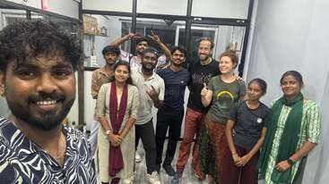 A group of ten people are standing together indoors, smiling for a selfie. The atmosphere looks friendly, casual, and diverse, with everyone posing happily together.