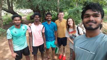 A group of six friends stands outdoors in a forested area, smiling at the camera. They are casually dressed, enjoying nature, and appear happy together.