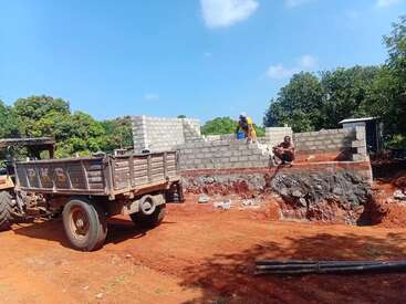 A construction site with two workers building a block wall, a tractor with a trailer, exposed soil, and green trees under a bright blue sky.