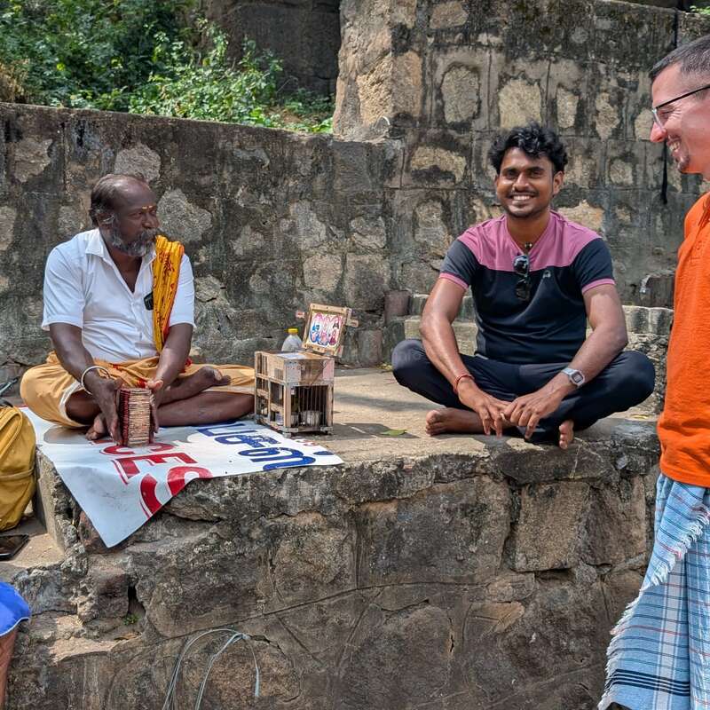 Four men sit and chat cheerfully outdoors against a stone wall. Two men wear traditional attire, while the other two wear casual clothes. Lively, friendly interaction.